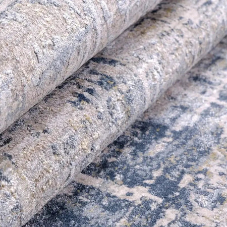 A close-up of a rolled area rug showing its textured, distressed pattern in beige, gray, and dark blue-gray tones.