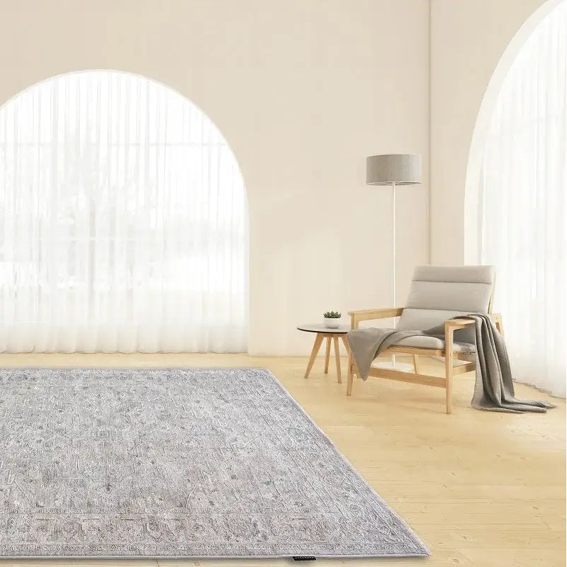 A rectangular area rug with a distressed, faded traditional pattern in shades of gray and white, displayed on a light wood floor in a bright, modern room with arched windows and a minimalist armchair.