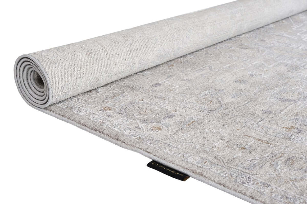 A light gray and beige patterned area rug, partially rolled up to show both its top surface and underside, photographed on a clean white background.