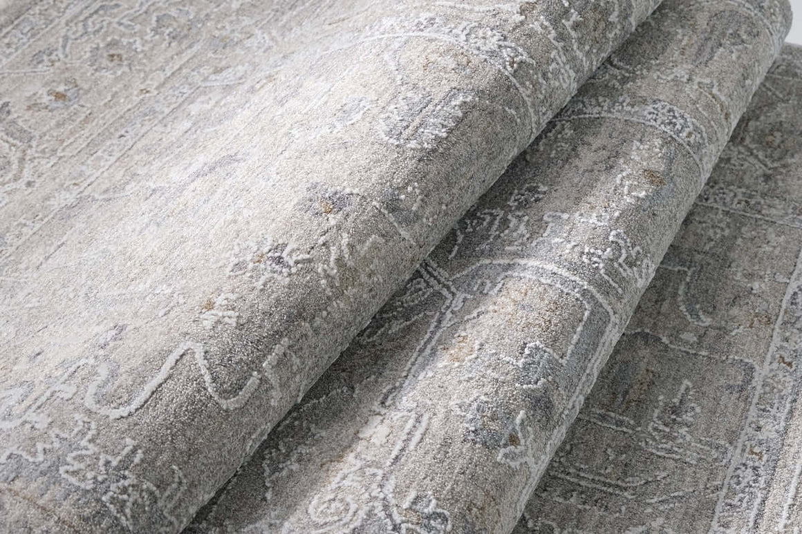 A close-up of a folded area rug with a distressed, traditional pattern in shades of gray, beige, and off-white.