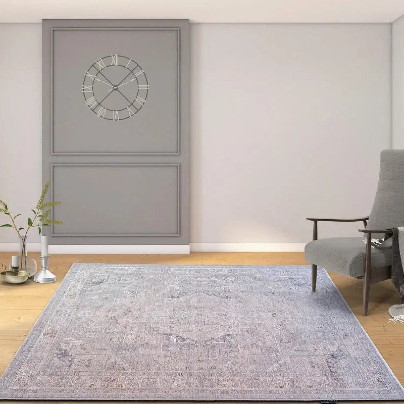 A large rectangular area rug with a faded blue and gray traditional pattern is displayed on a light wood floor in a modern room setting. The room features light gray walls, a gray accent wall with a circular clock, a gray armchair, and a small side table.
