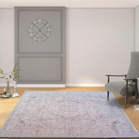A large rectangular area rug with a faded blue and gray traditional pattern is displayed on a light wood floor in a modern room setting. The room features light gray walls, a gray accent wall with a circular clock, a gray armchair, and a small side table.