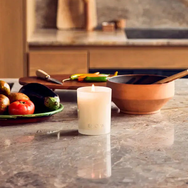 Candle on a kitchen counter with vegetables and a cutting board in the background