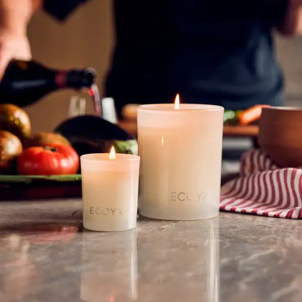 Two lit candles on a kitchen counter with a person and kitchen items in the background.