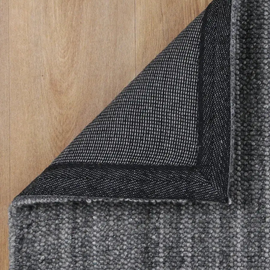 Close-up of a dark gray textured rug with one corner folded back to show the backing, placed on a light wood floor.
