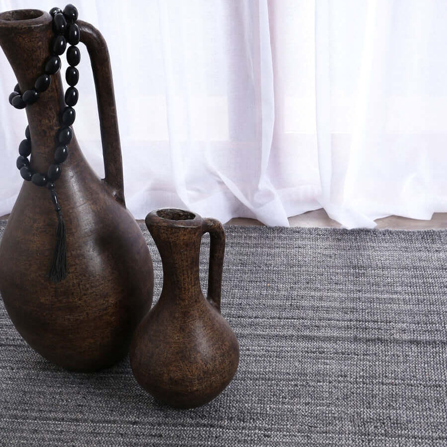 A dark gray woven area rug with a textured horizontal ribbed pattern, styled in a room setting with two rustic dark brown ceramic pitchers and a white curtain in the background.