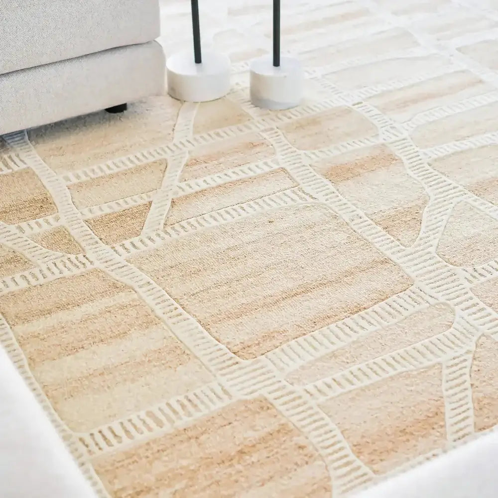 A close-up of a light beige and cream area rug with a geometric block pattern, styled in a modern living room setting with a sofa corner and two floor lamp bases visible.