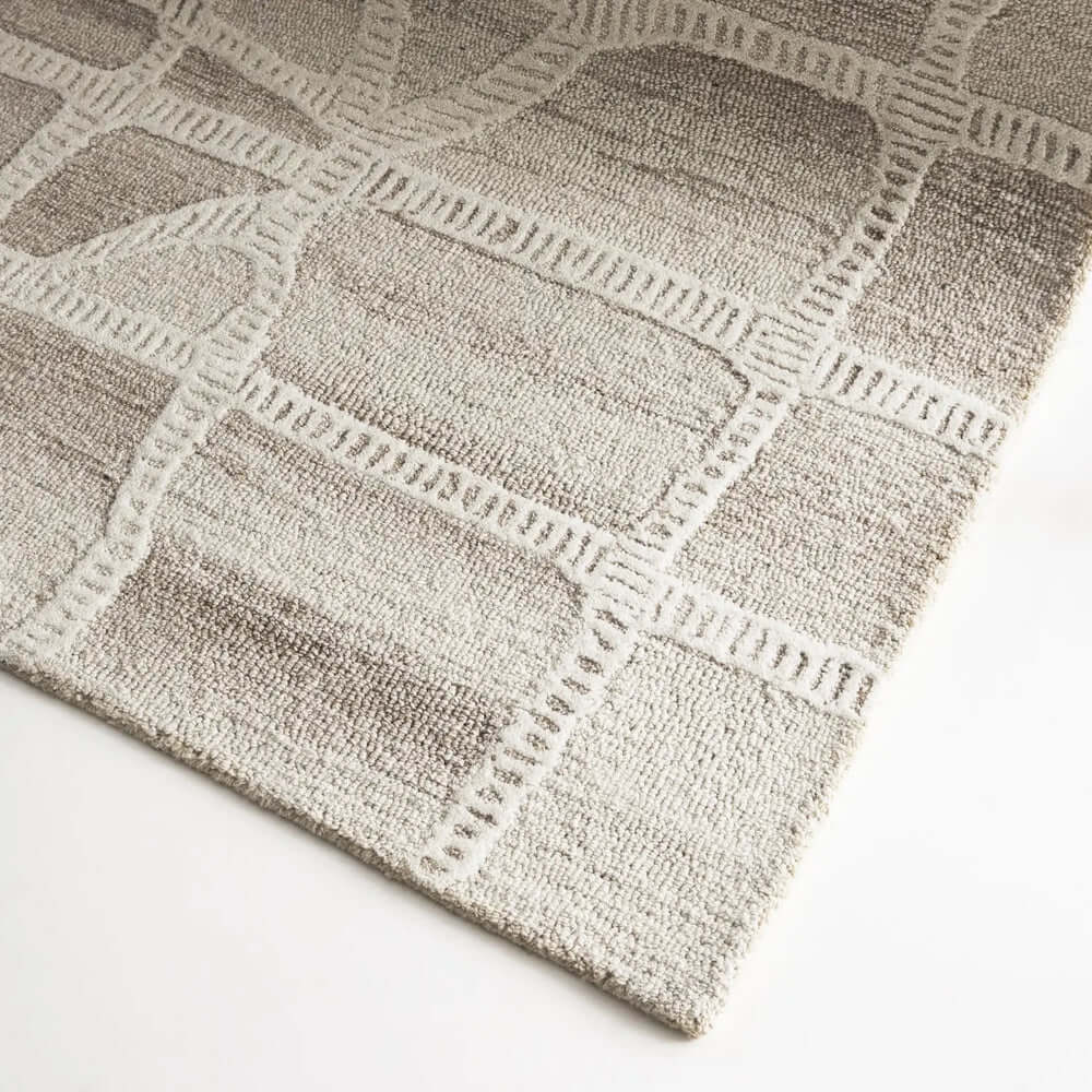 A close-up of a textured area rug with a geometric lattice pattern in shades of taupe, beige, and off-white, shown on a clean white surface.