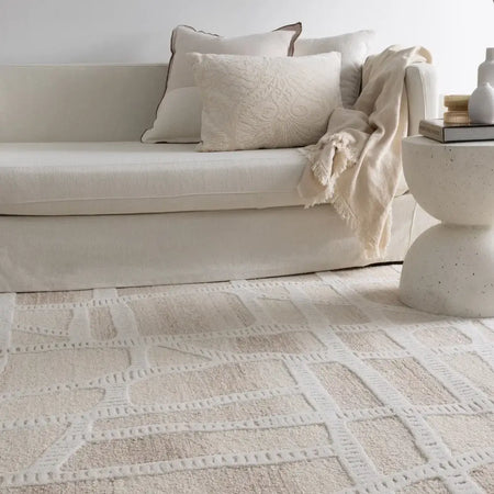 A low-profile upholstered sofa in a warm off-white fabric, styled with neutral throw pillows and a cream knit throw blanket, set in a bright room on a textured cream and beige patterned area rug. A matching textured side table sits to the right.